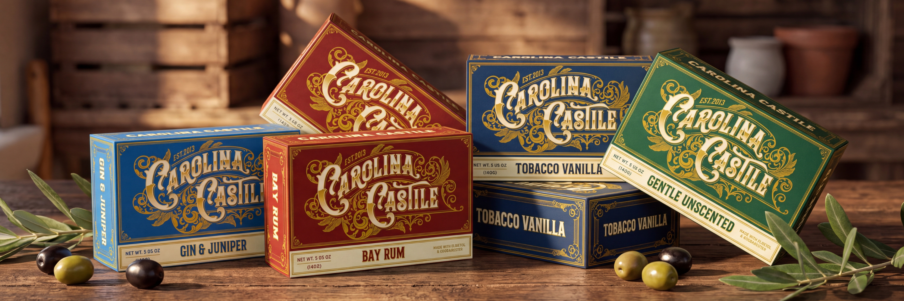 Five Carolina Cabin tobacco product boxes arranged on a rustic wooden table with olives and leaves in the foreground. The boxes are shown in blue, red, and green with gold lettering, and the background features a warm, blurred interior with wooden barrels.