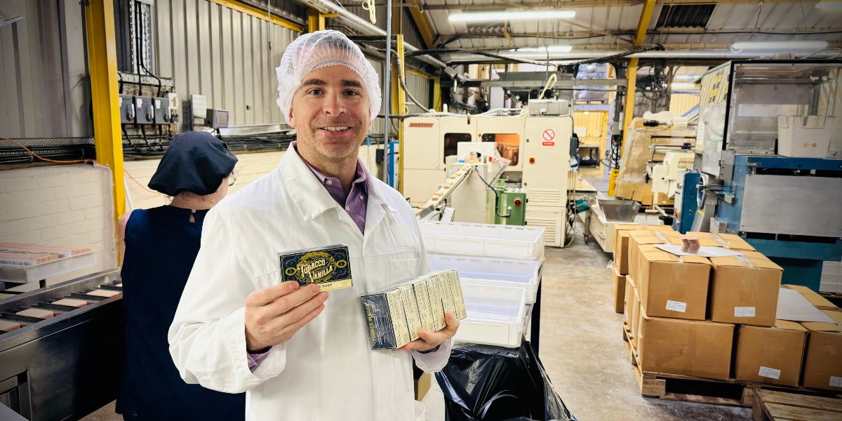 Joe McCutcheon holding Tobacco Vanilla Bar soap while standing in front of bar soap manufacturing line with conveyor carrying bars of soap