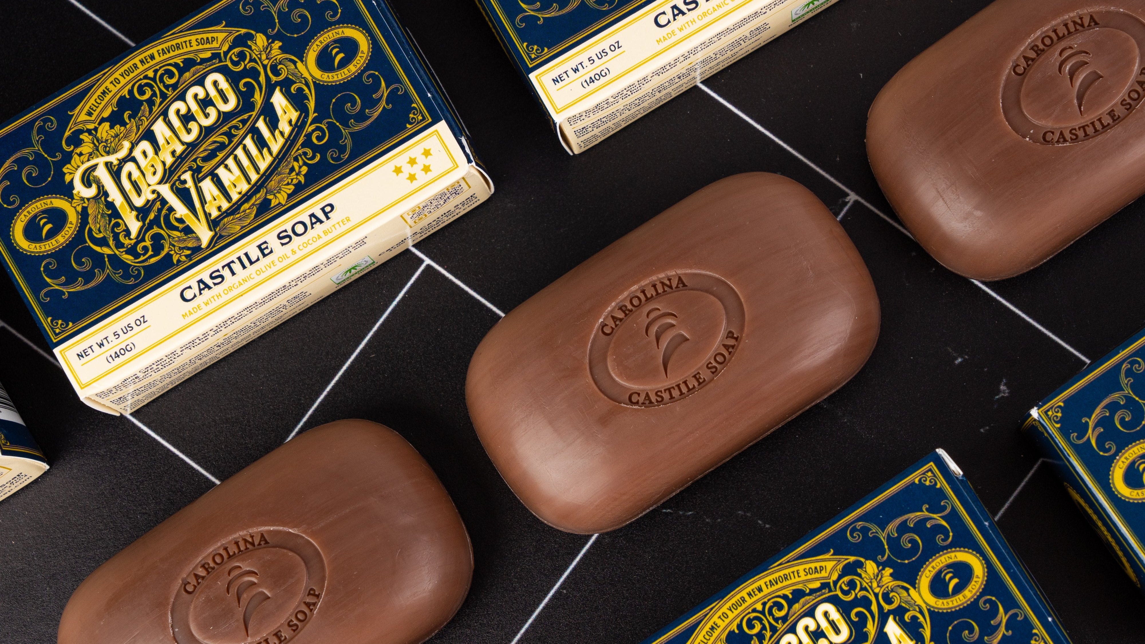 Arrangement of brown, Tobacco Vanilla bar soap with the logo for Carolina Castile Soap stamped into the bars. The bars are dark brown and are surrounded by the outer cartons for the bars which are graphical and have blue. All is arranged against a backdrop of black tile.